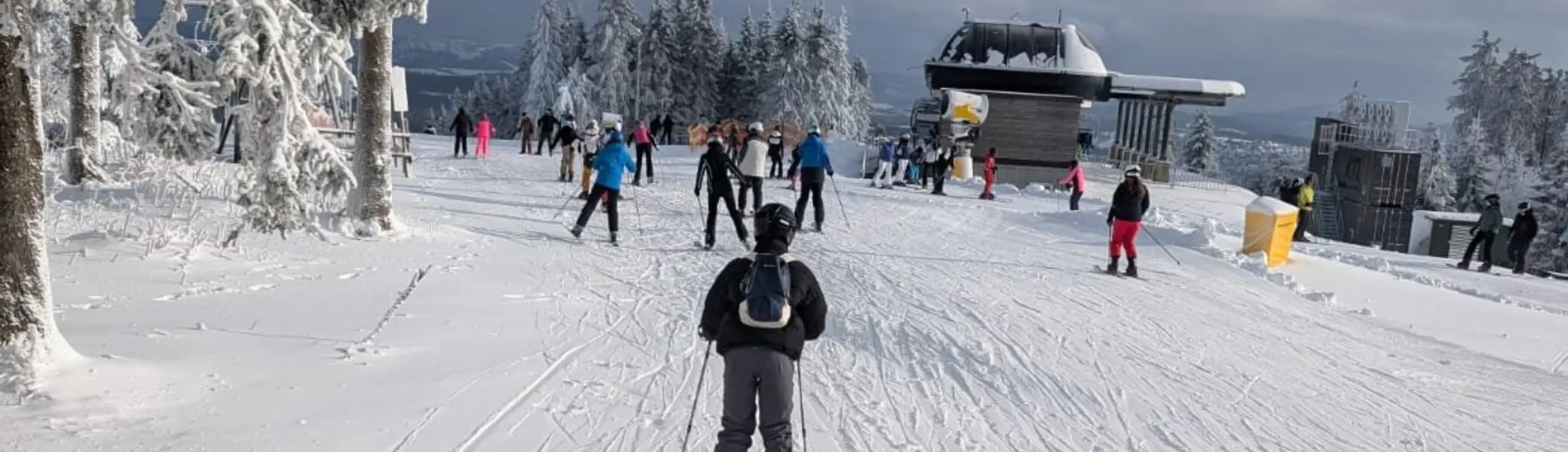 Einzelne und mehrere Skifahrer*innen auf breiter, sonniger Skipiste bei Bergstation. | © Dav Dortmund