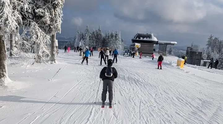 Einzelne und mehrere Skifahrer*innen auf breiter, sonniger Skipiste bei Bergstation. | © Dav Dortmund