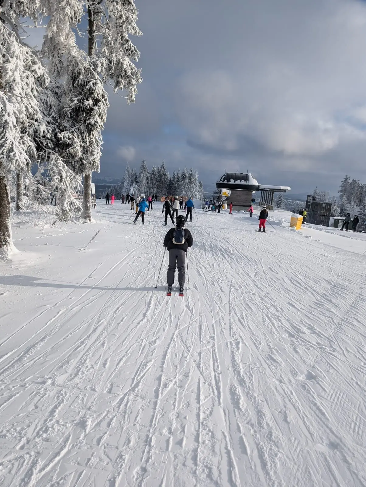 Einzelne und mehrere Skifahrer*innen auf breiter, sonniger Skipiste bei Bergstation. | © Dav Dortmund