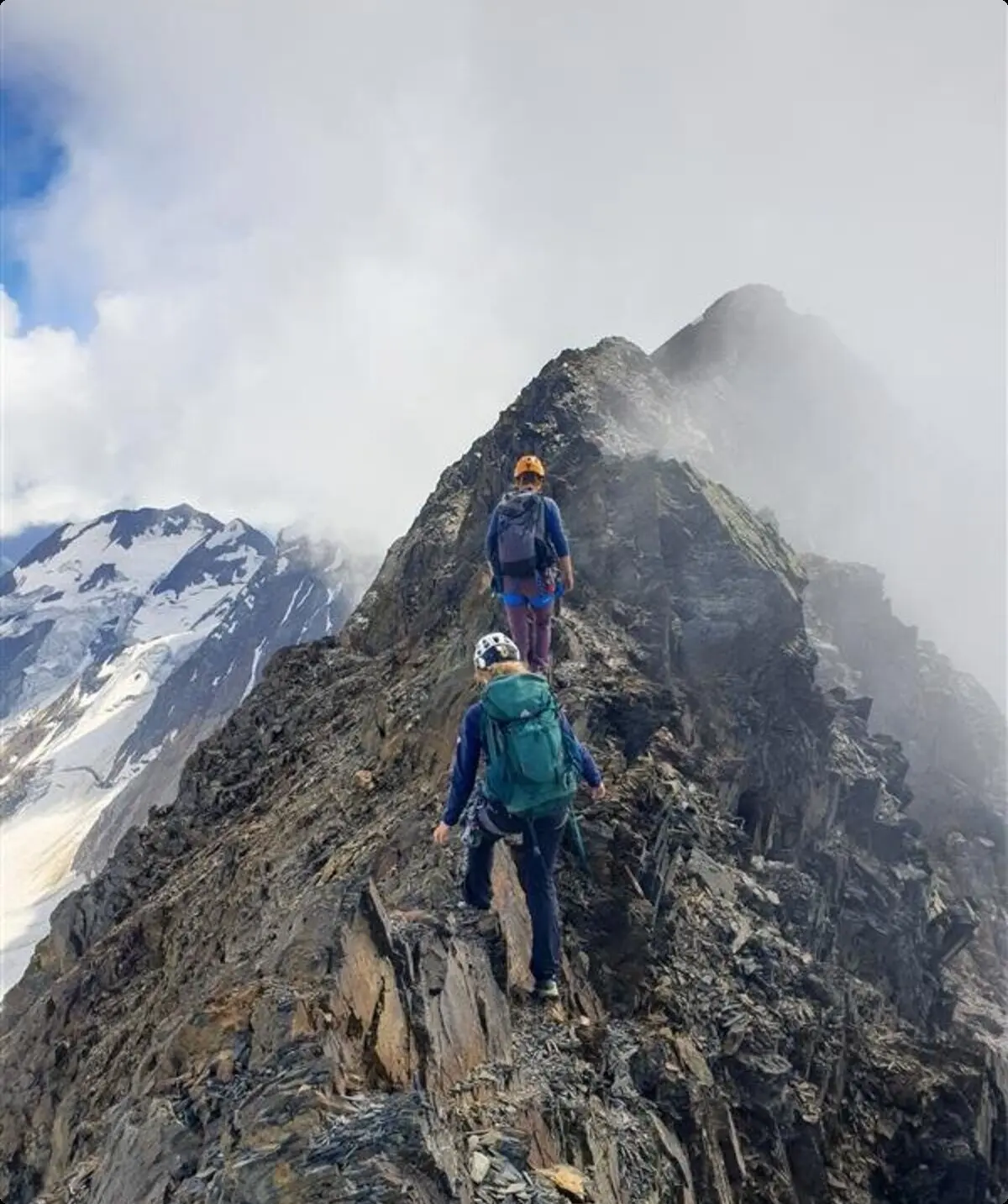 Zwei Bergsteiger*innen gehen über einen schmalen, felsigen Grat mit Blick auf Gletscher und Wolken. | © jdav nrw