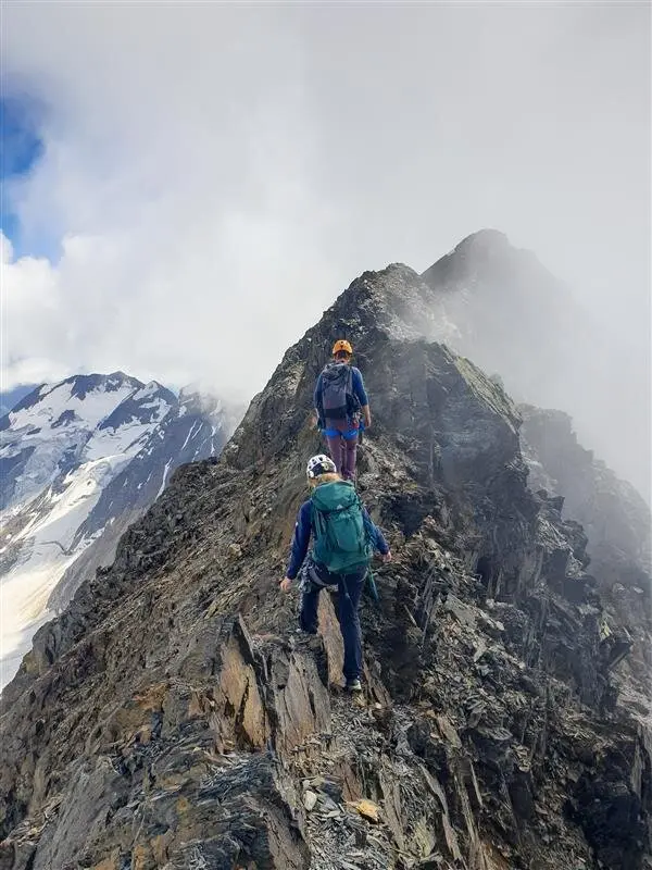 Zwei Bergsteiger*innen gehen über einen schmalen, felsigen Grat mit Blick auf Gletscher und Wolken. | © jdav nrw