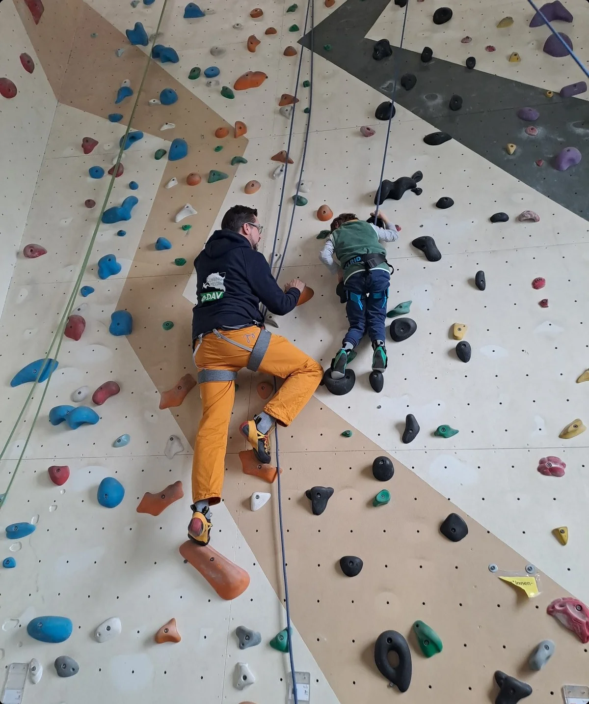 Ein Erwachsener sichert ein Kind beim Klettern an einer Indoorwand mit bunten Griffen. | © DAV Dortmund