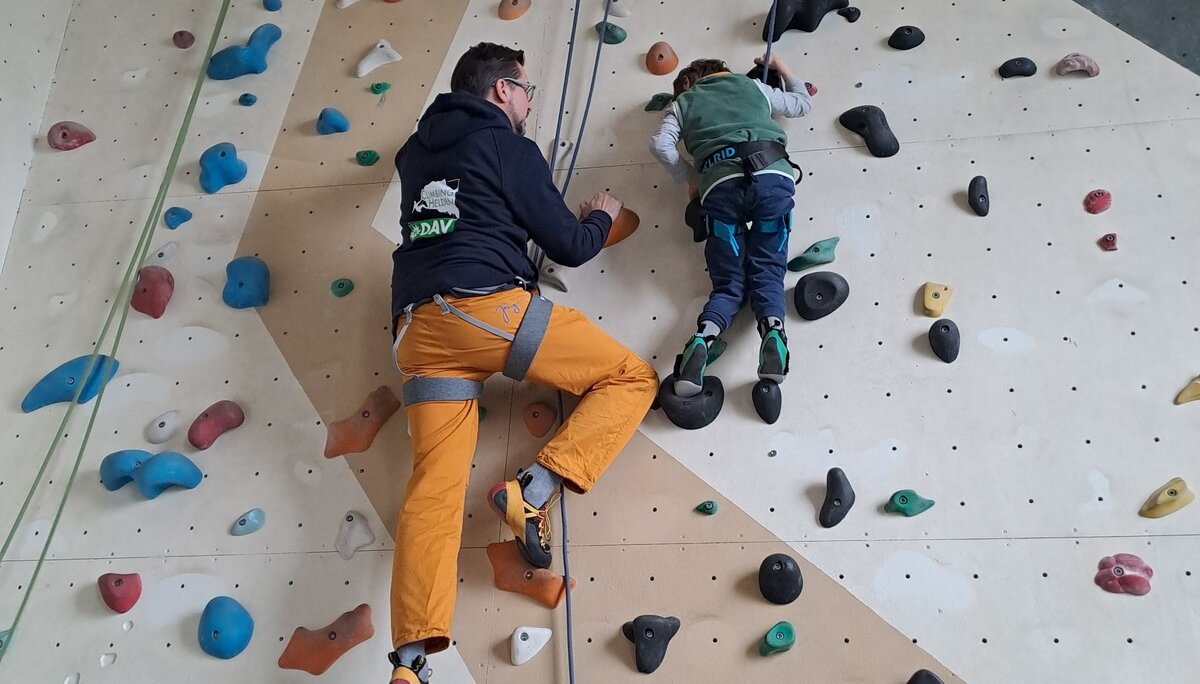 Ein Erwachsener sichert ein Kind beim Klettern an einer Indoorwand mit bunten Griffen. | © DAV Dortmund