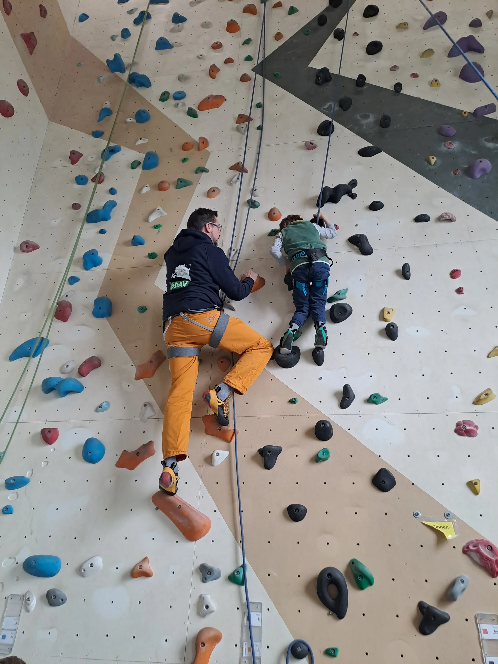 Ein Erwachsener sichert ein Kind beim Klettern an einer Indoorwand mit bunten Griffen. | © DAV Dortmund