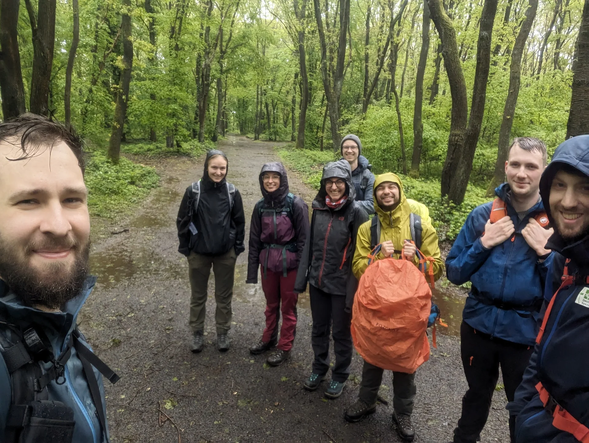 Acht Personen stehen mit Regenjacken auf einem Waldweg und lächeln in die Kamera. | © u27 - JDAV