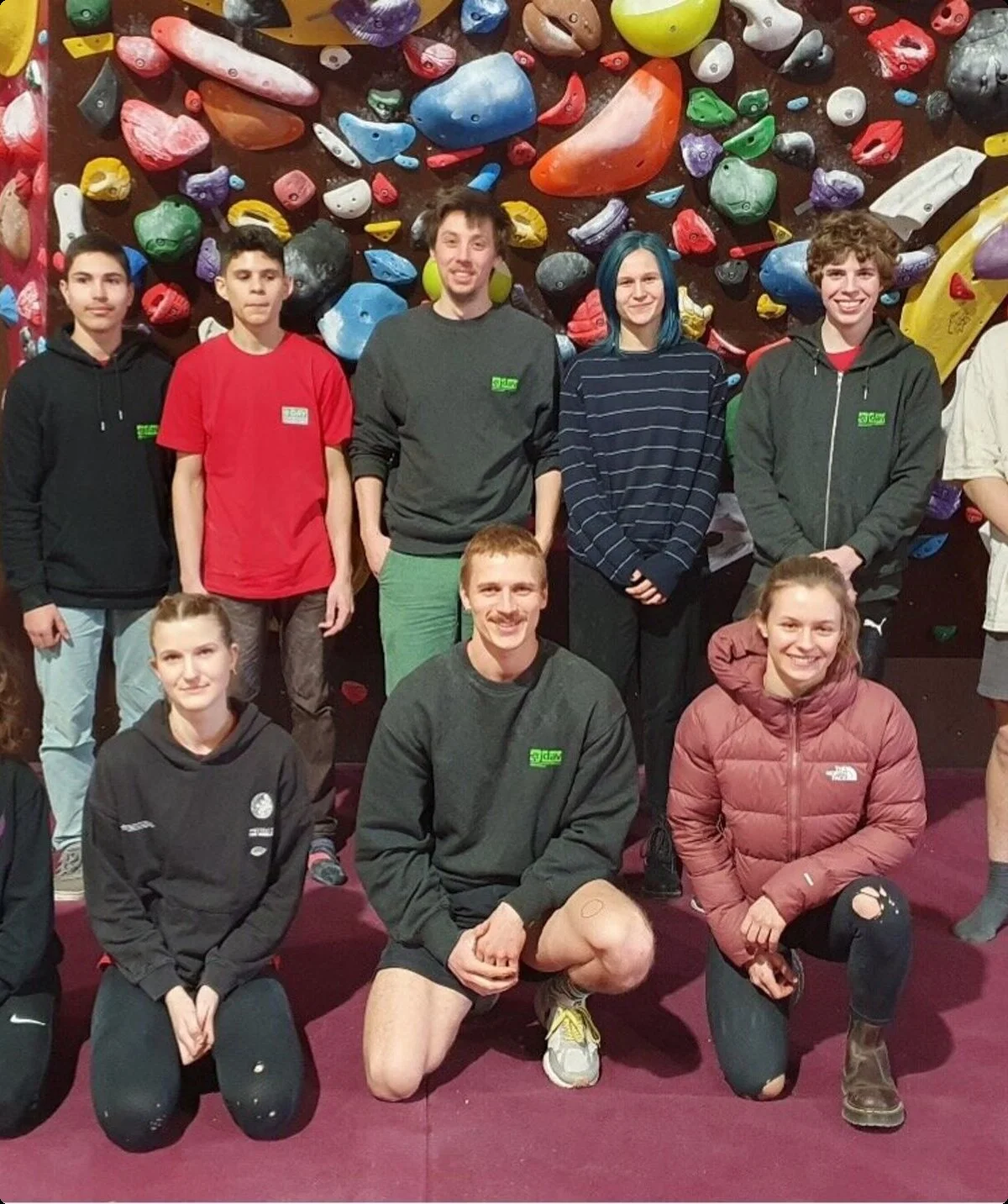 Jugendliche und junge Erwachsene stehen und knien vor einer bunten Boulderwand auf roter Matte. | © DAV Dortmund