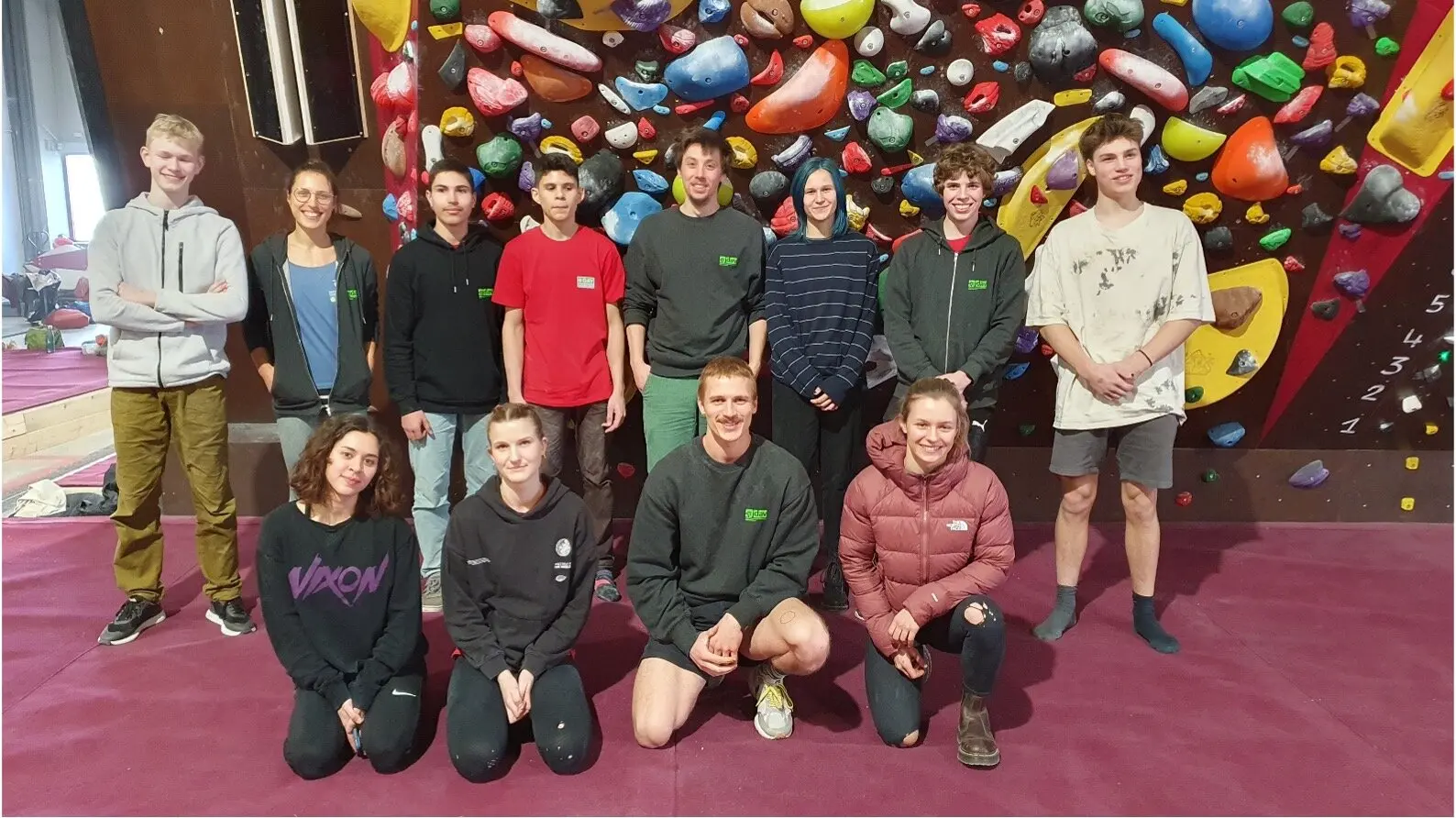Jugendliche und junge Erwachsene stehen und knien vor einer bunten Boulderwand auf roter Matte. | © DAV Dortmund
