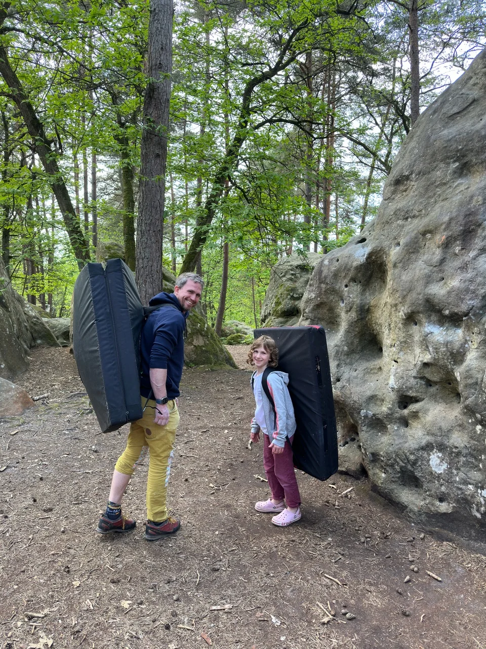 Ein Erwachsener und ein Kind mit Crashpads auf dem Rücken beim Bouldern im Wald. | © DAV Dortmund